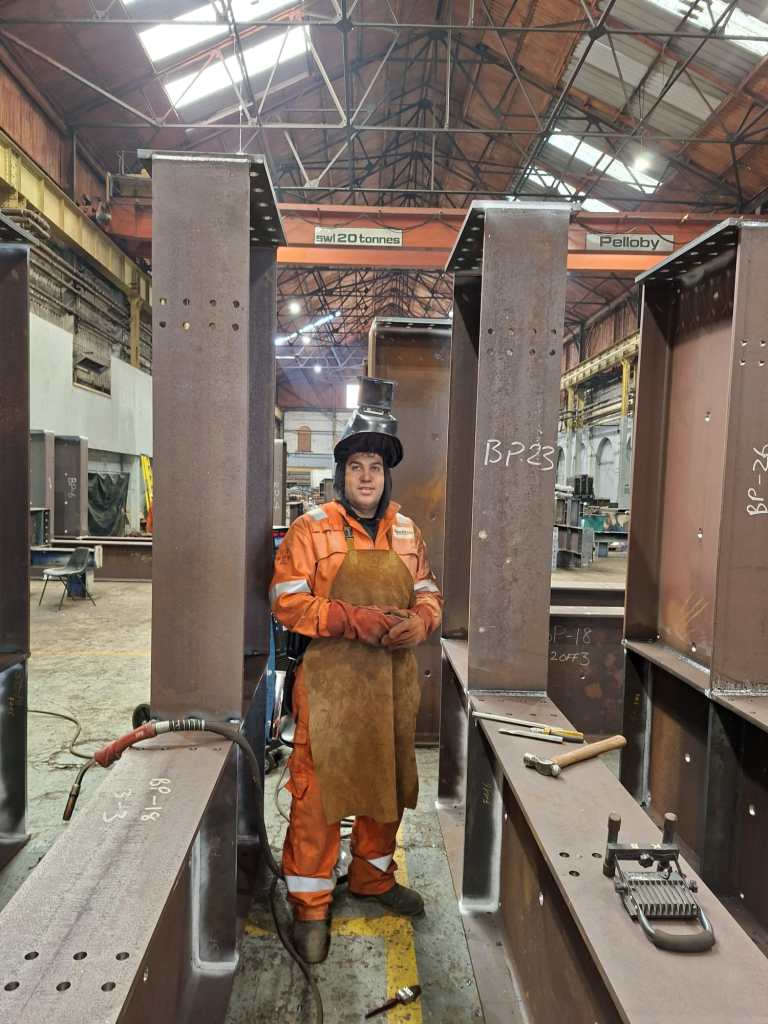 Welder in orange hi-vis standing between steel columns in workshop