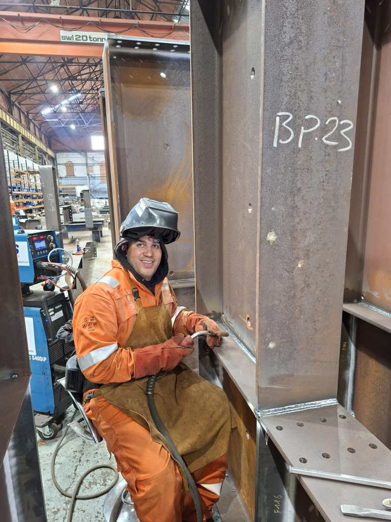 Welder in orange hi-vis smiling while welding a steel column
