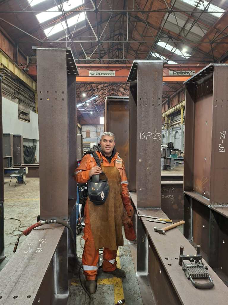 Welder in orange hi-vis standing between steel columns in workshop